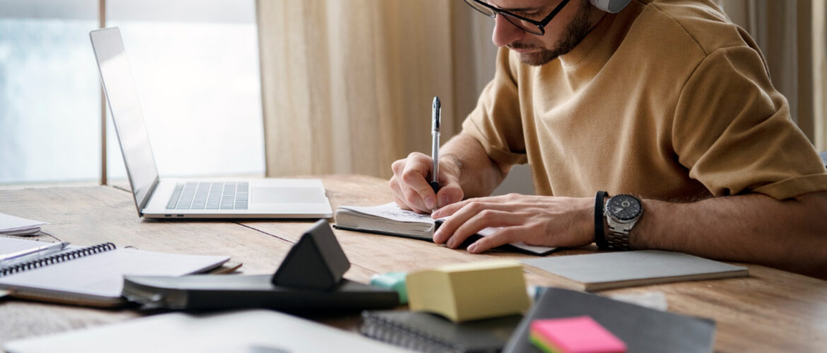 A man with headphones writes in a notebook at a desk with a laptop and stationery.