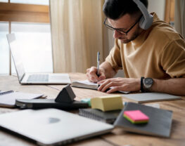 A man with headphones writes in a notebook at a desk with a laptop and stationery.