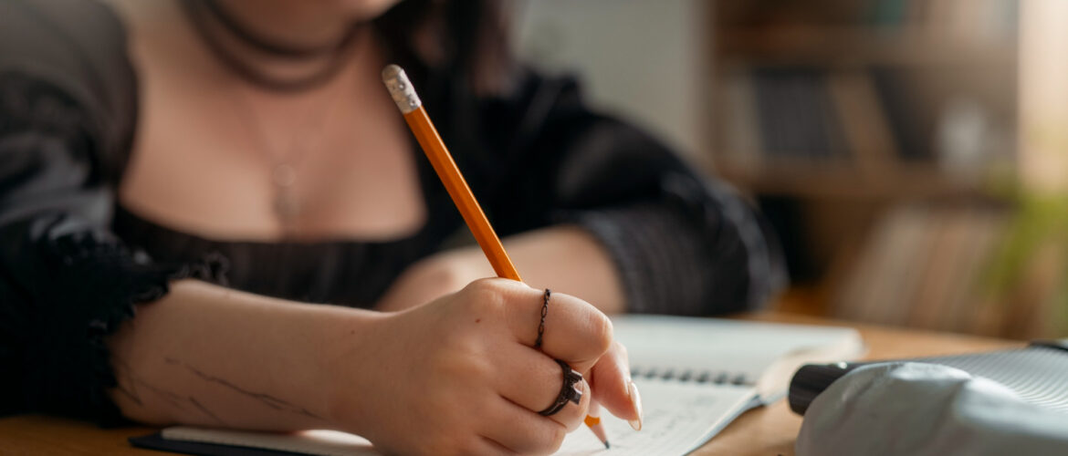 A close-up of a gothic-style girl writing in a notebook, wearing dark clothing and rings, with blurred bookshelves in the background.