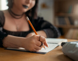 A close-up of a gothic-style girl writing in a notebook, wearing dark clothing and rings, with blurred bookshelves in the background.